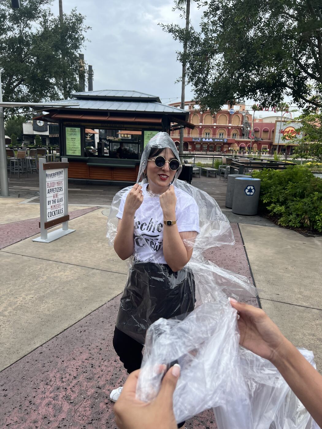 Karishma in round sunglasses wrapped in a clear plastic rain poncho, fists up, on an outdoor plaza.