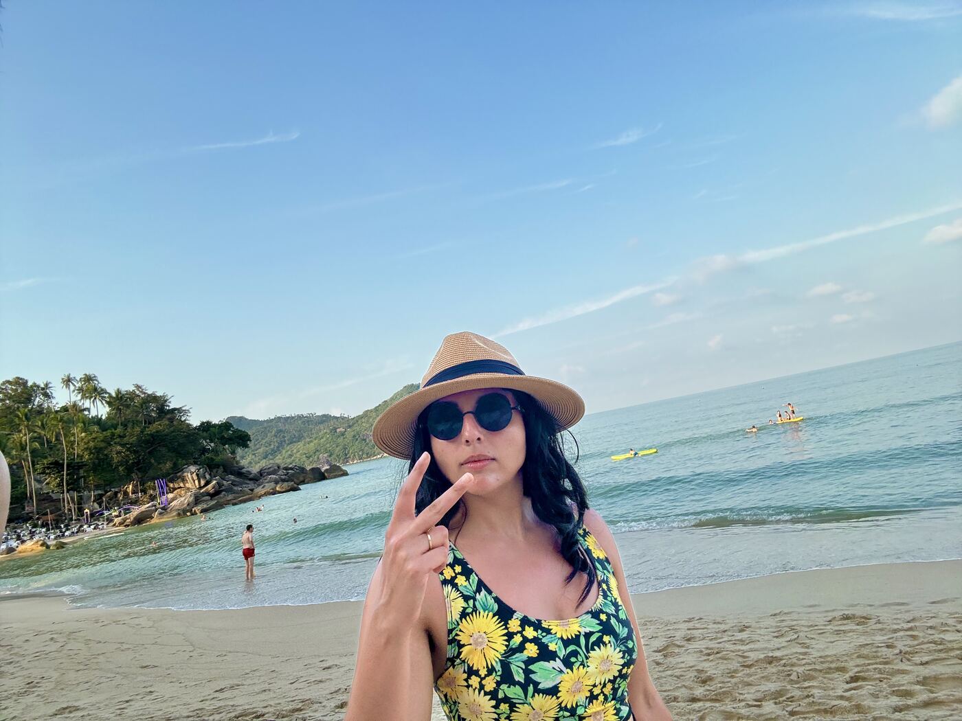 Karishma on a beach in a straw hat, round sunglasses and a sunflower print swimsuit, holding up a peace sign.